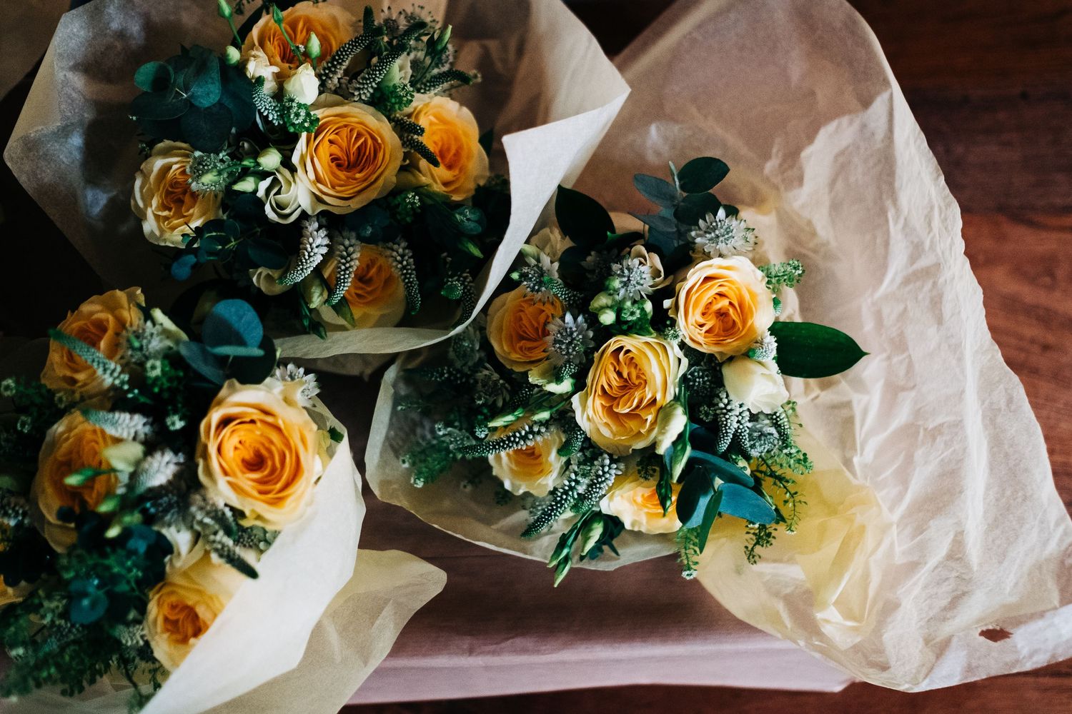 Three elegant bouquets of peach-colored roses wrapped in white paper sit on a wooden surface.