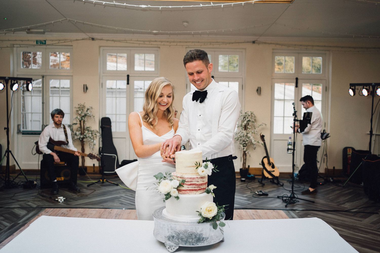 Elmhay Park Wedding Photos Newlyweds cut their elegant white wedding cake together while a band sets up in the background at their reception