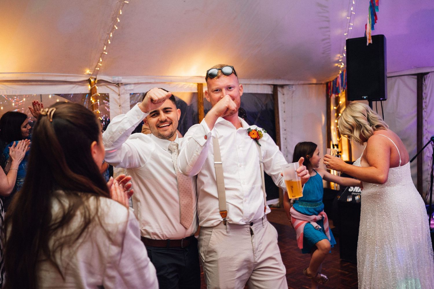 Wedding guests dance and celebrate together on a dimly lit dance floor with purple lighting effects.