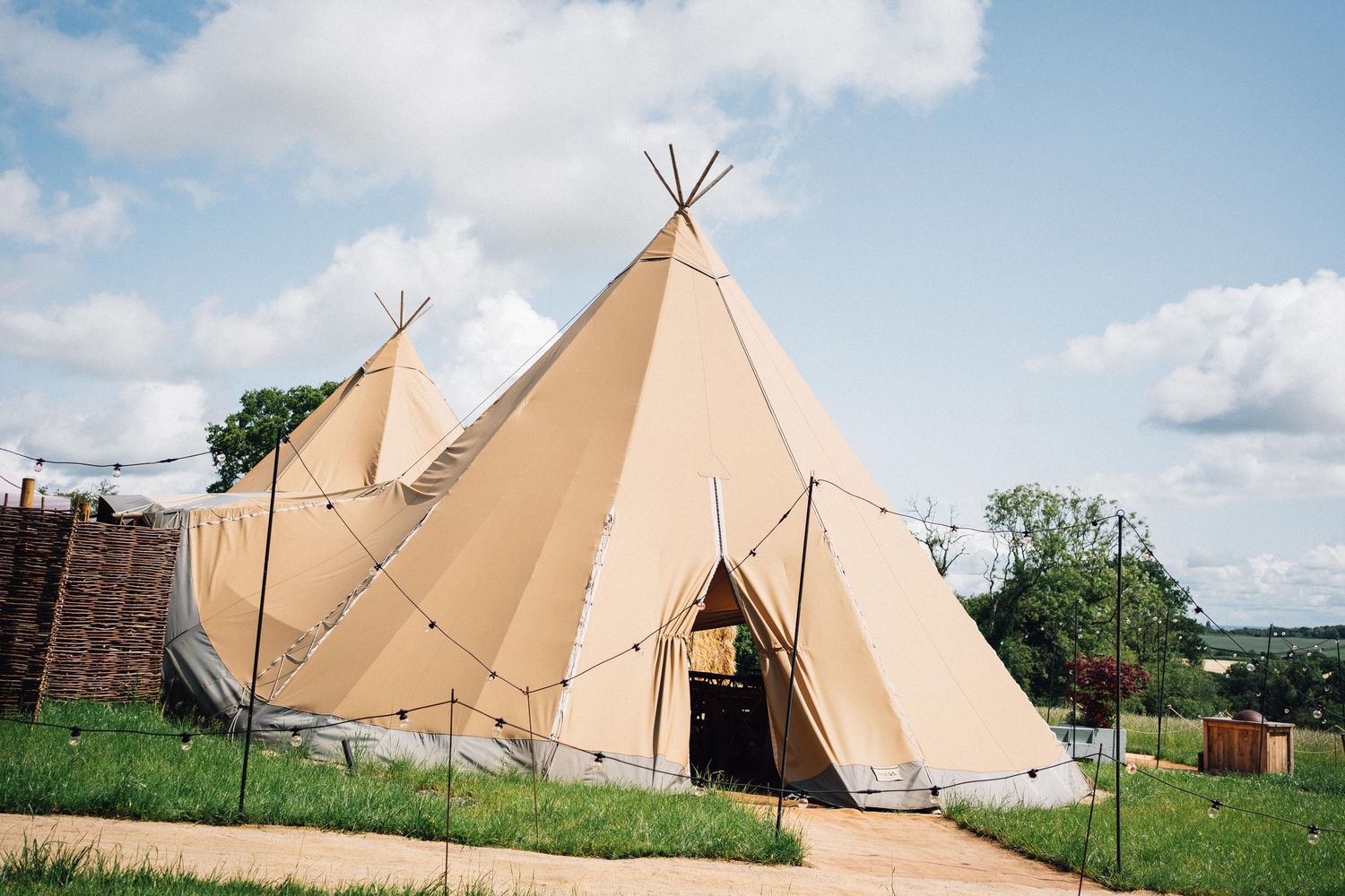 Two large beige teepee tents set up in a grassy field under a blue cloudy sky.