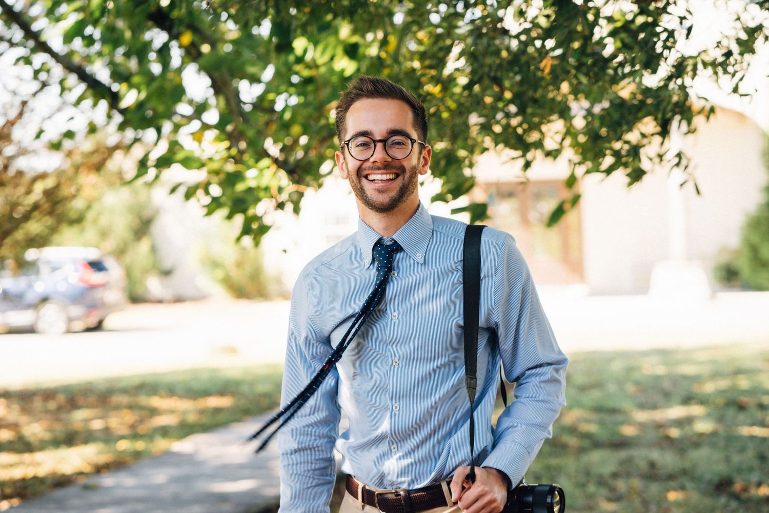 Professional wearing a light blue button-down shirt smiles while sitting outdoors in a natural setting.