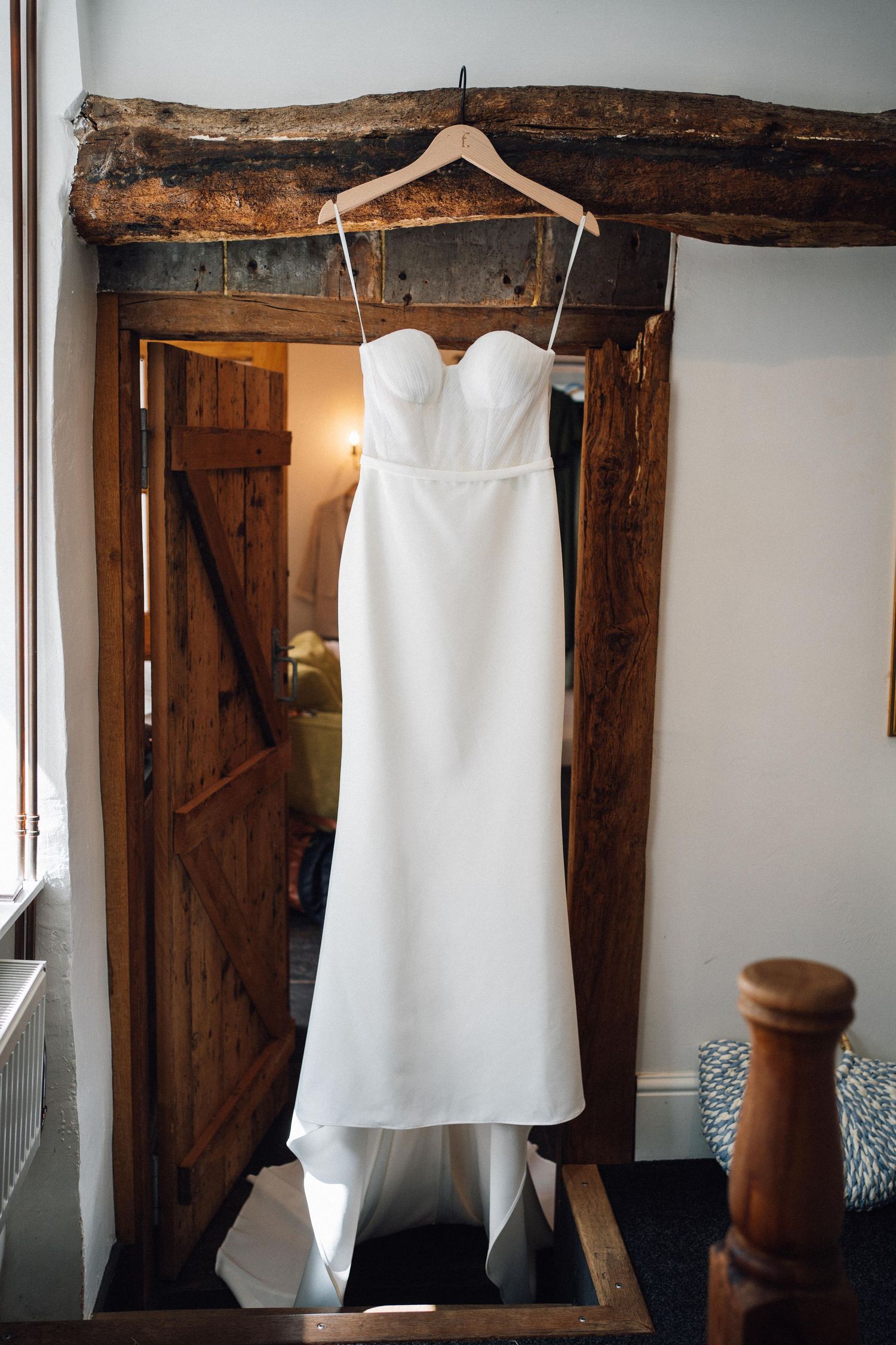 White wedding dress hangs from rustic wooden beams in a vintage room with natural lighting.