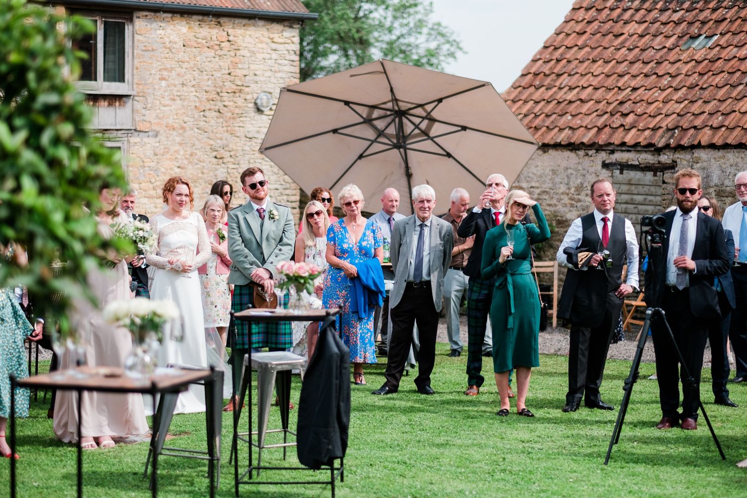 Wedding guests gather on a lush green lawn outside a rustic stone building for an outdoor reception celebration.