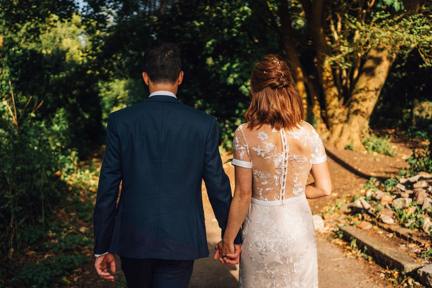 Newlyweds walking through sunlit garden path surrounded by lush greenery.