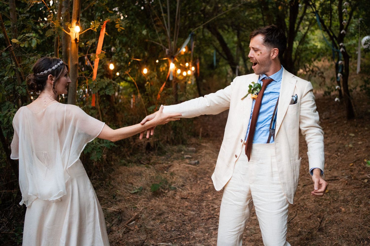 A bride and groom hold hands and smile at each other in a romantic forest wedding setting.