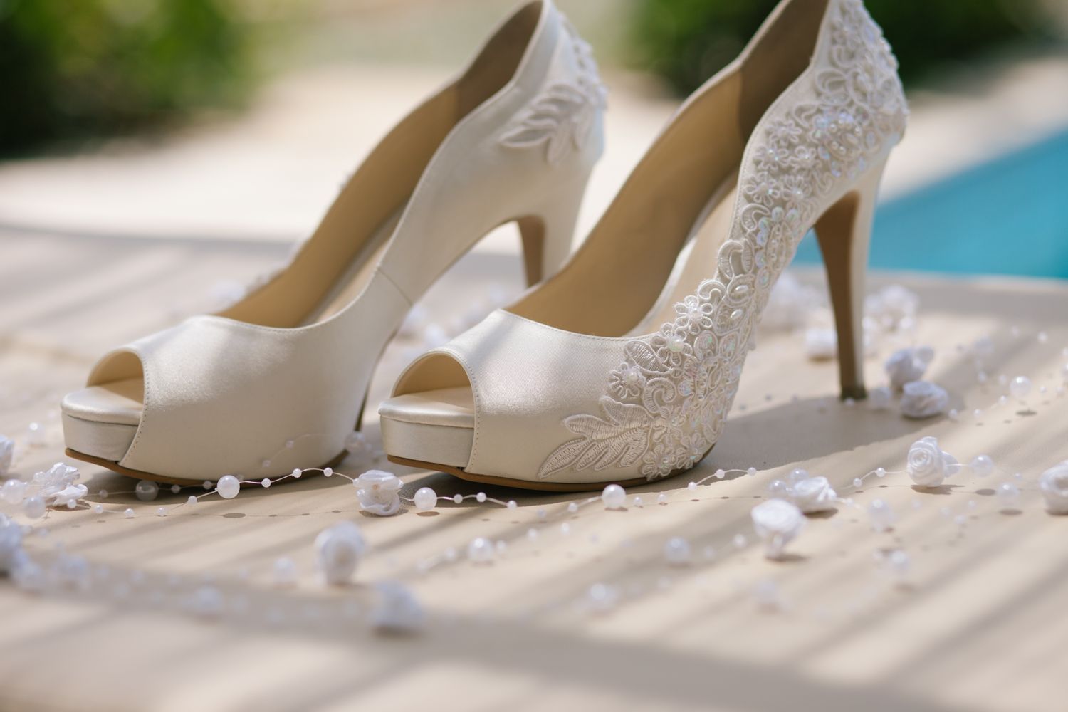 Elegant white lace wedding shoes displayed on a sandy beach with ocean and palm trees in background.