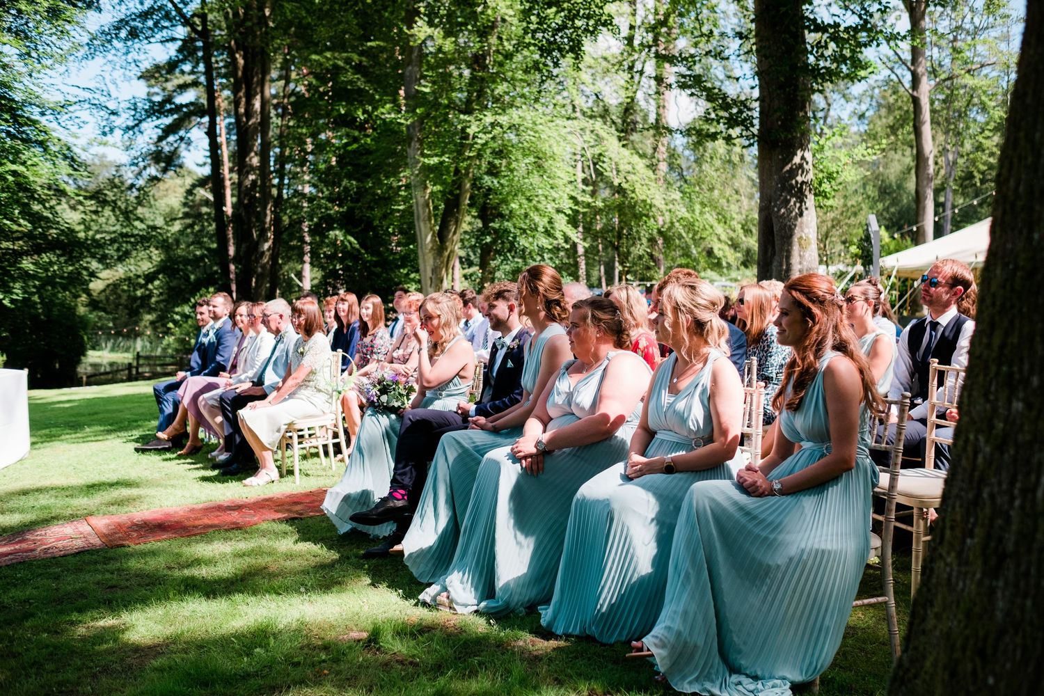 Wedding guests seated in rows of white chairs during an outdoor forest ceremony.