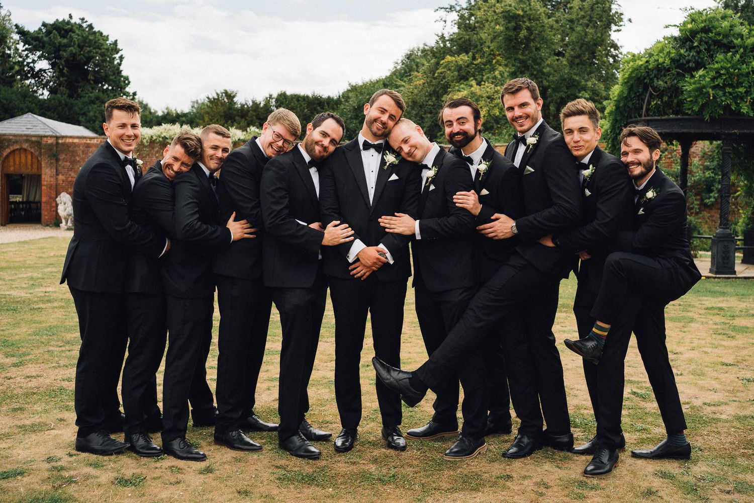 Group of groomsmen in formal attire striking fun poses outdoors at wedding venue.