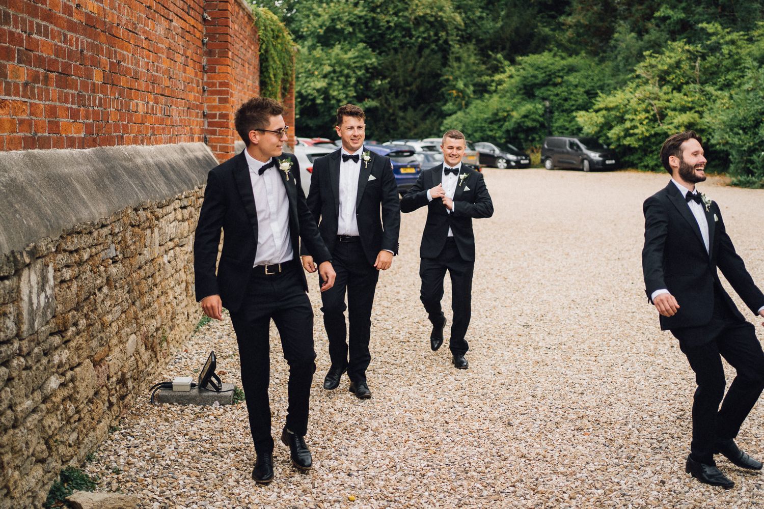 Groomsmen in formal black tuxedos gather on a gravel path near brick walls.