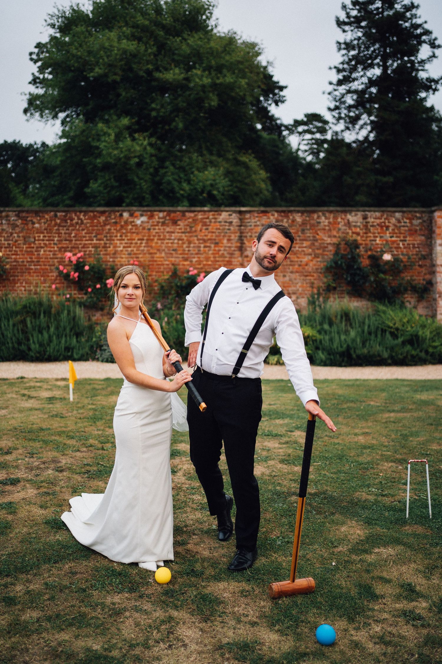 Newlyweds play croquet on a lawn with brick wall backdrop during wedding reception.