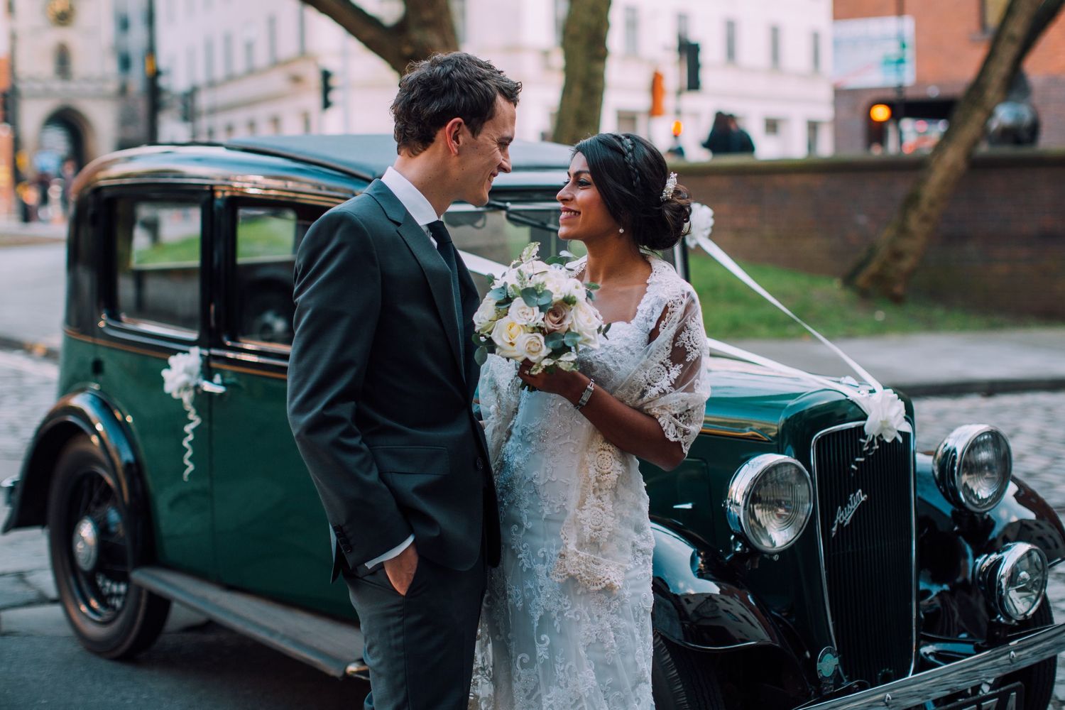 Bride and groom pose with vintage green car during wedding photoshoot at sunset.