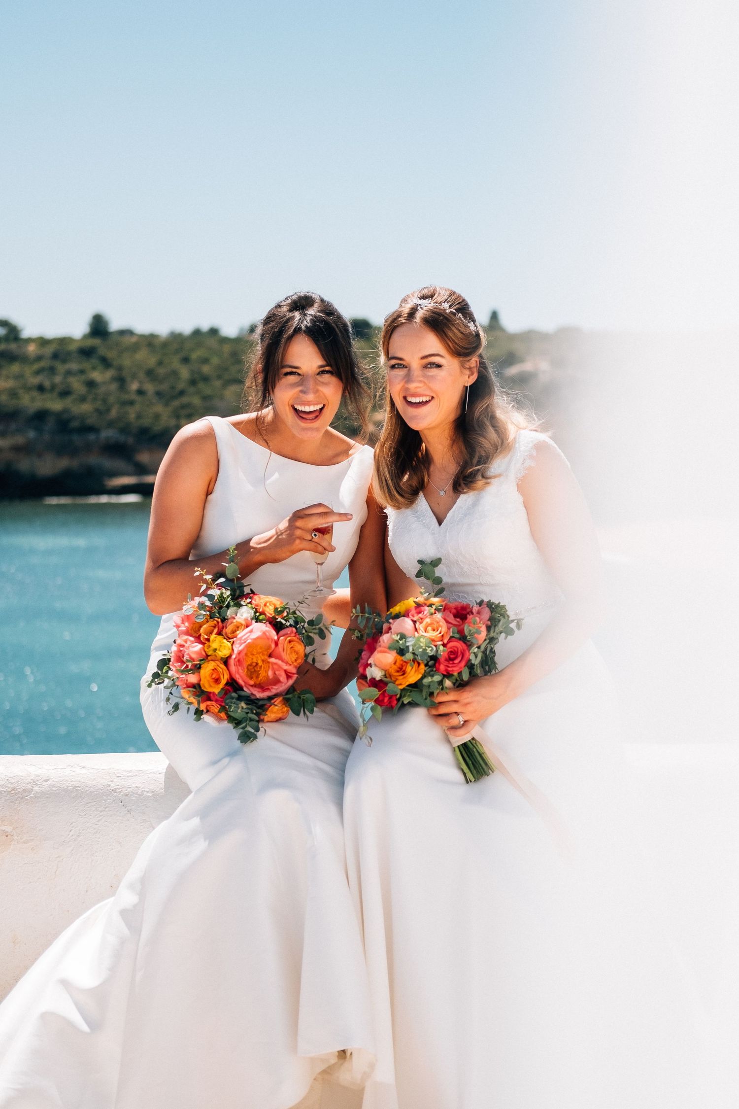 Two people in white wedding dresses laugh together while holding vibrant orange and pink floral bouquets by the water.