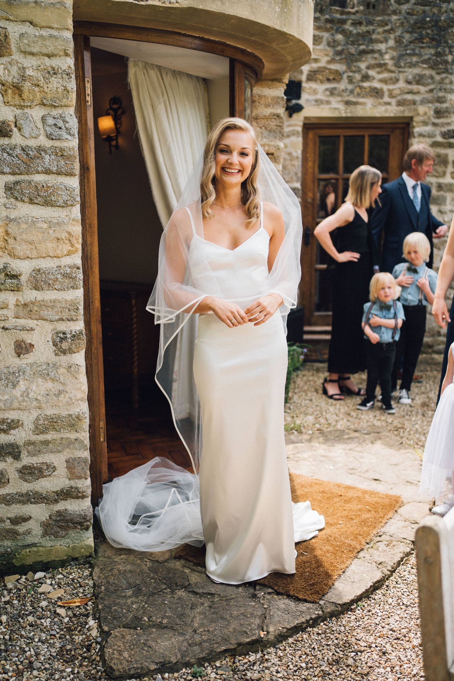 A bride in a flowing white wedding dress sits gracefully on stone steps outside a rustic building with stone walls.
