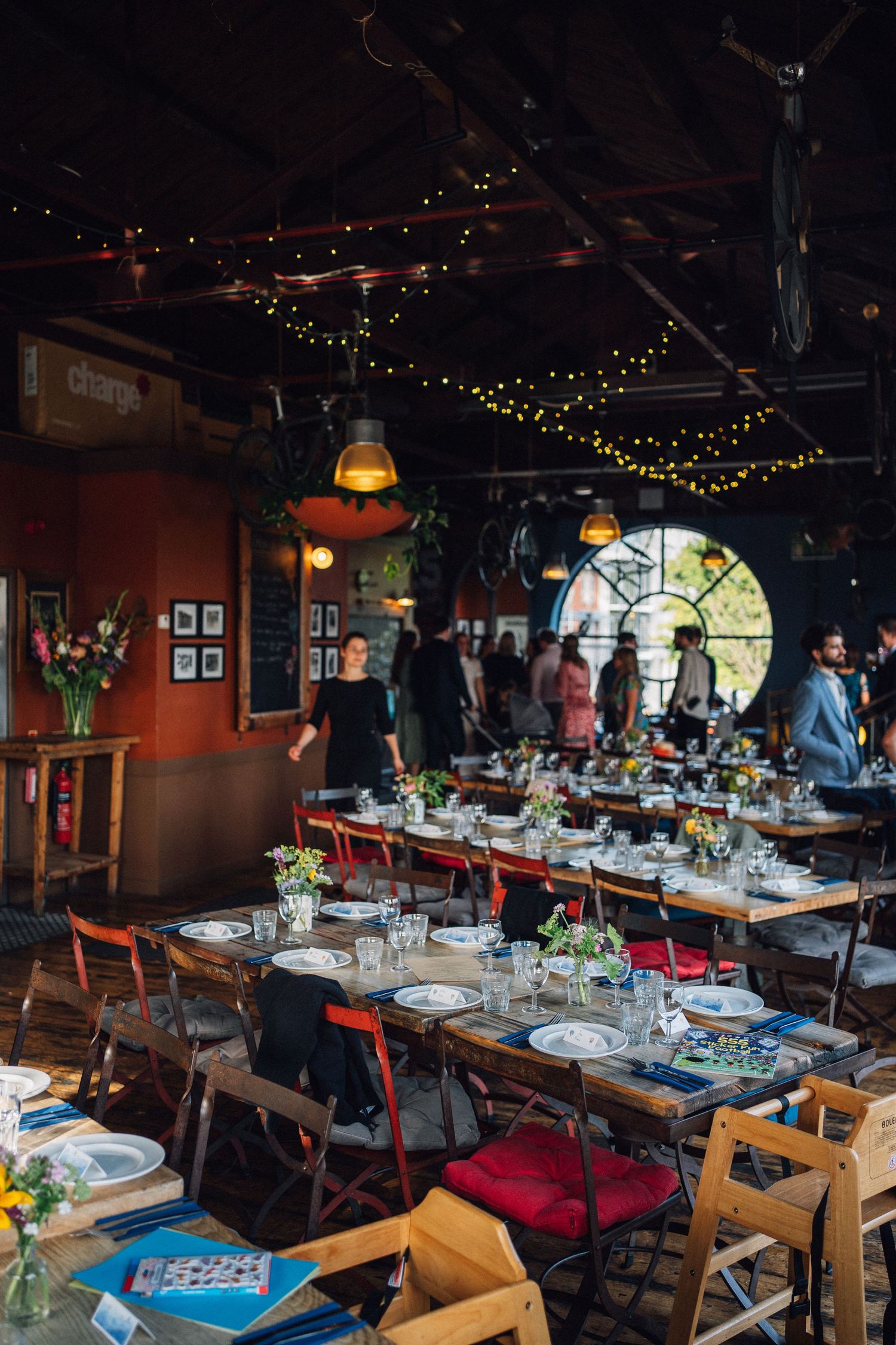 Rustic event venue with long wooden tables set for dinner, string lights, and guests mingling in the background.