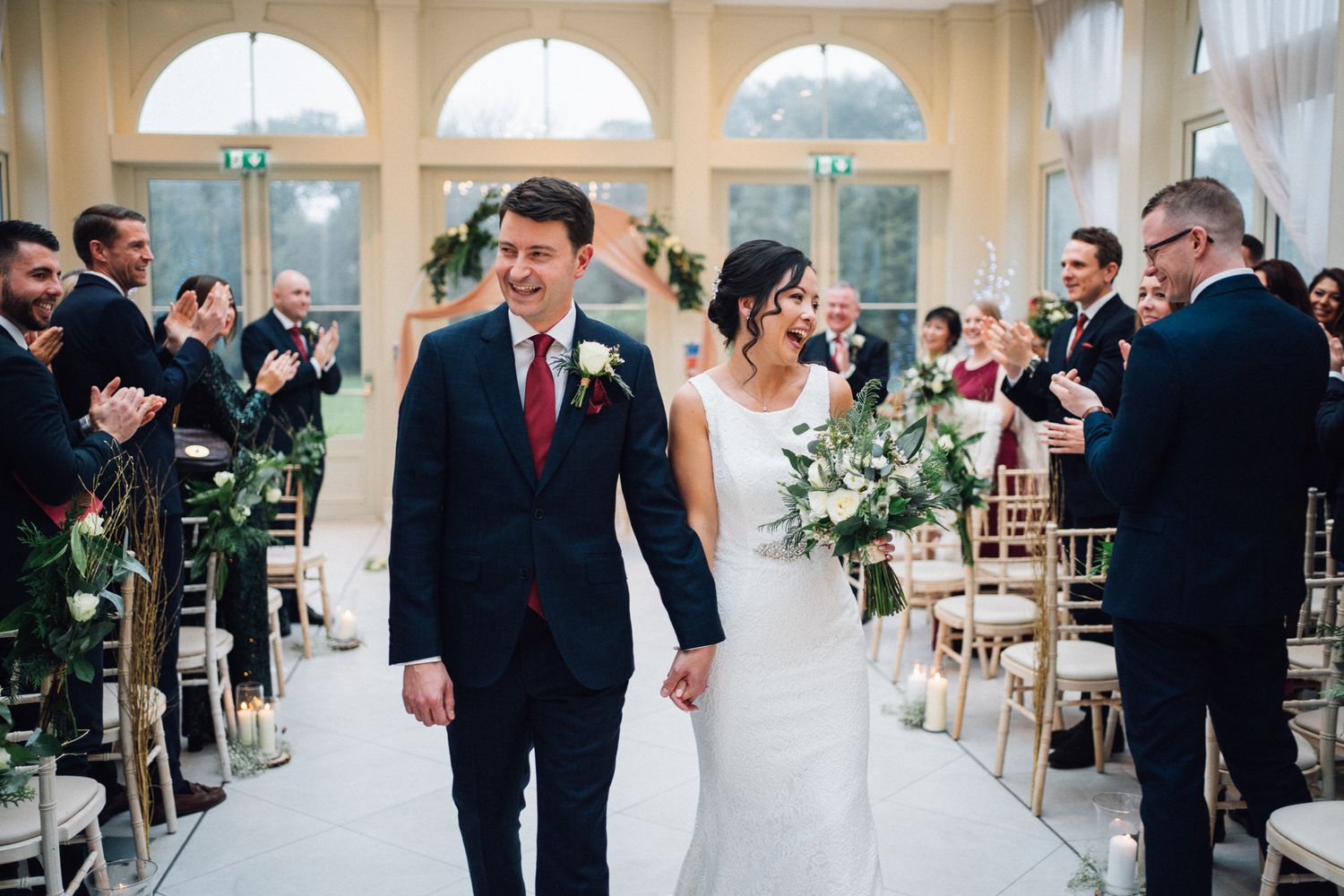Bride and groom walk down the aisle smiling at their elegant indoor wedding ceremony with guests applauding.