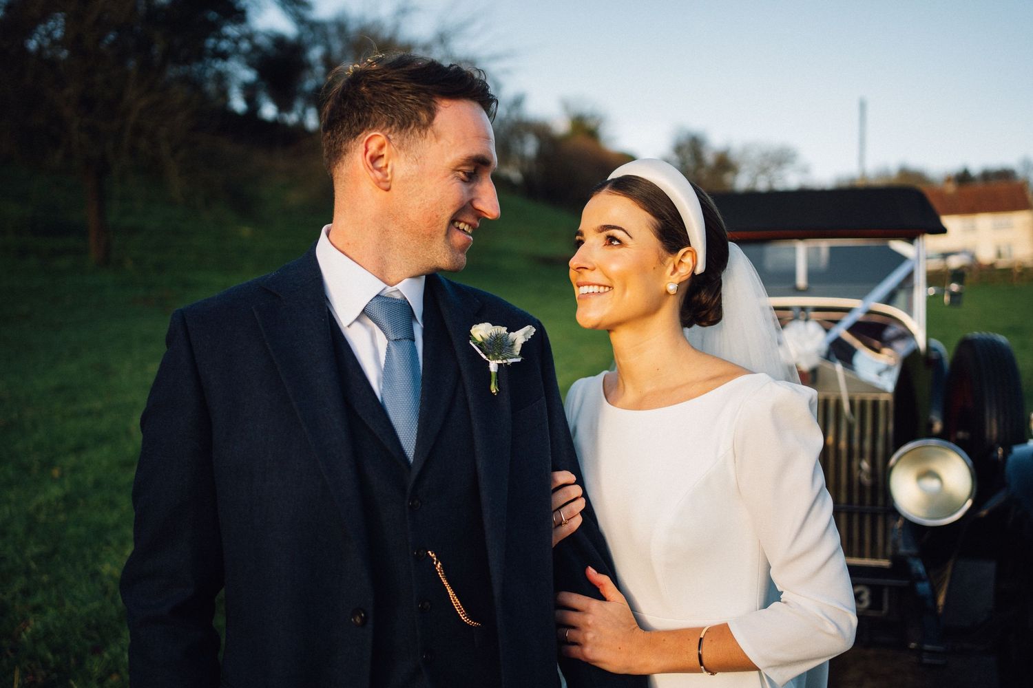 A smiling bride and groom in wedding attire stand together outdoors near a vintage car on their wedding day.