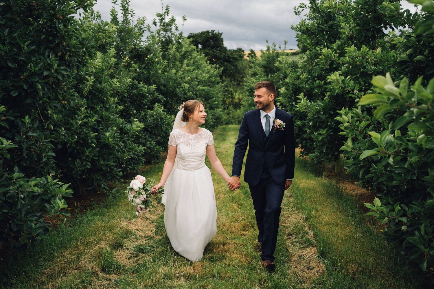 Couple walking through lush green orchard rows on their wedding day.