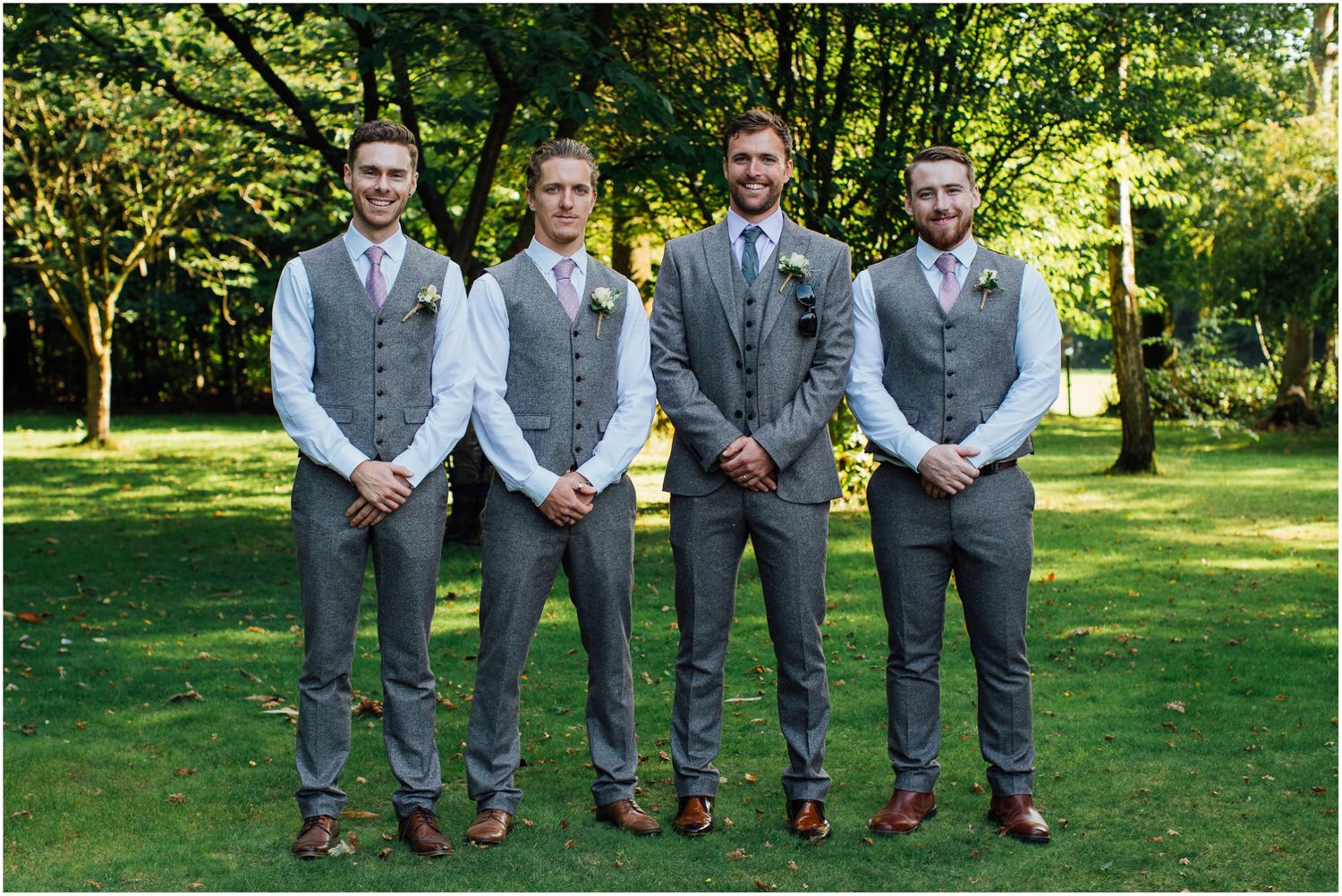 Groomsmen in matching grey suits stand together on green lawn.