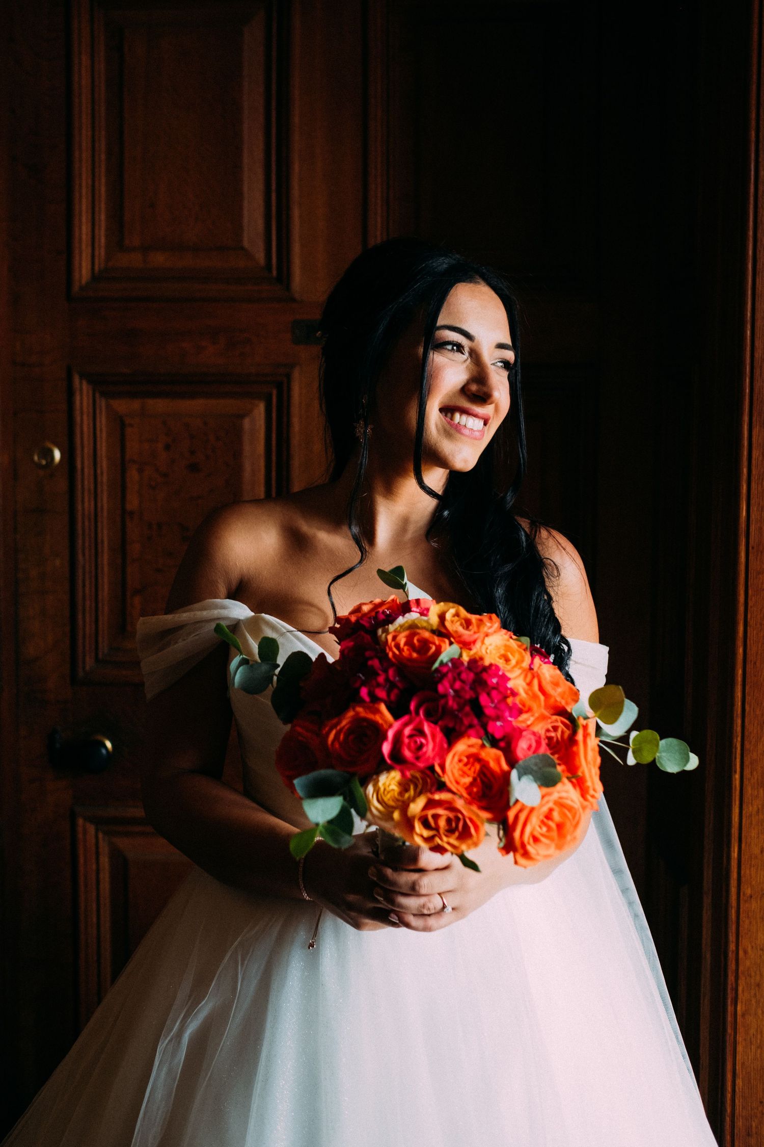 Dramatic lighting illuminates a bridal bouquet of orange roses against a white gown.