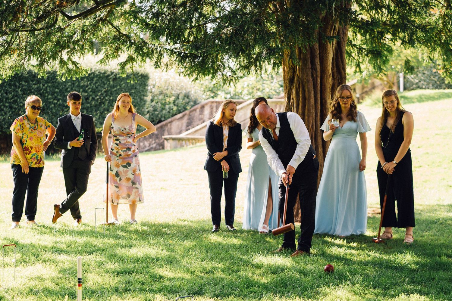 Wedding party and guests gathered for photos under green trees in a garden setting.