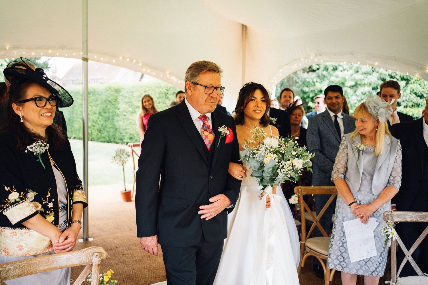 Wedding guests gather under a white tent for an outdoor celebration with chairs arranged in rows.