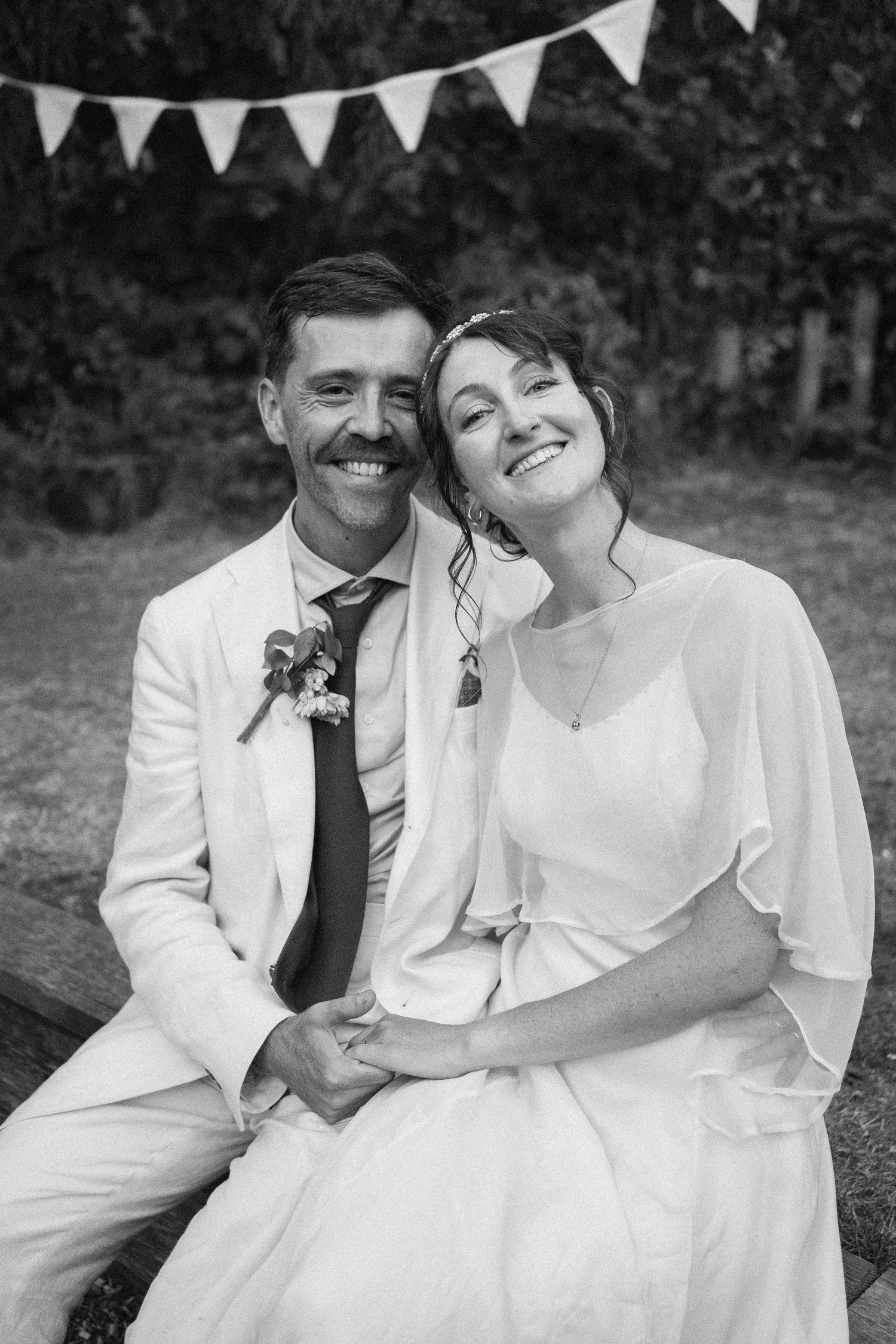 Black and white portrait of smiling bride and groom sitting together with bunting in background.