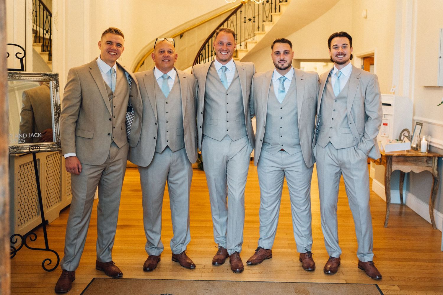 Five groomsmen in light blue suits pose together in a rustic venue hallway.