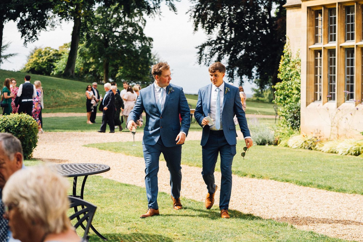 Two guests in blue suits walk together on the lawn of an elegant country estate venue.
