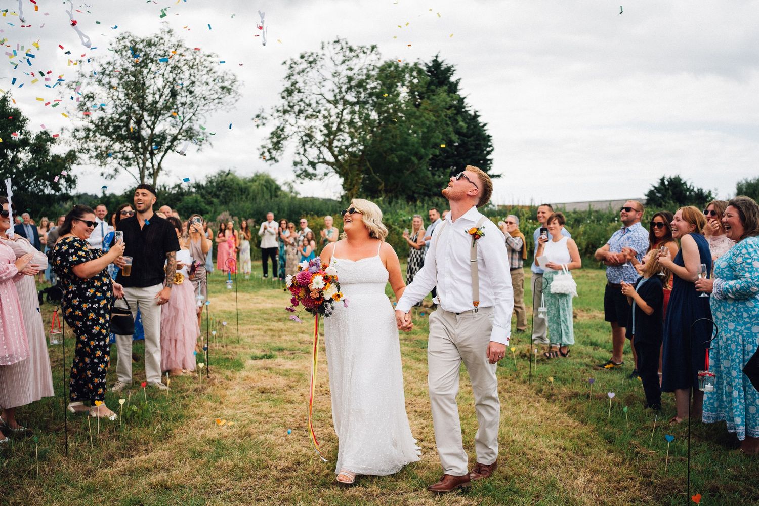 Wedding guests shower confetti over newlyweds walking down the aisle at an outdoor summer ceremony.