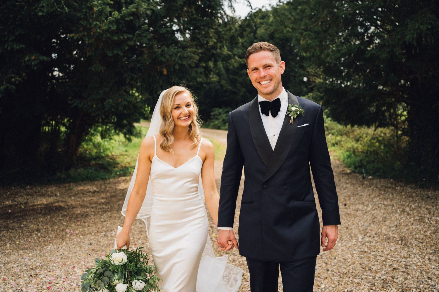 Bride and groom holding hands smiling on gravel path surrounded by trees, bride in white gown, groom in navy tuxedo.
