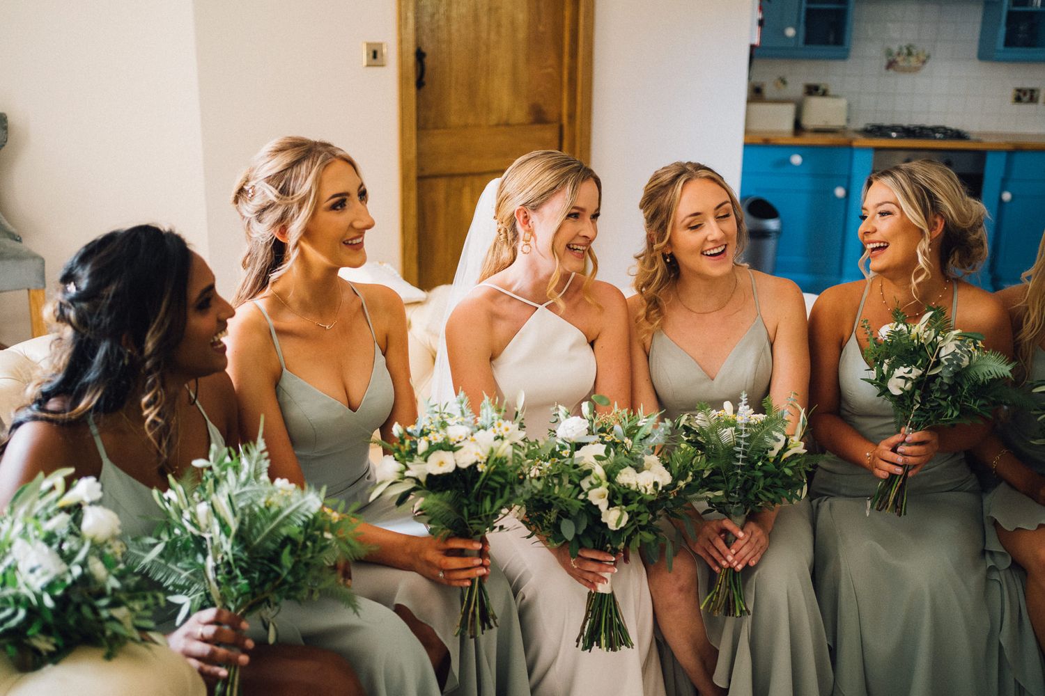 Row of bridesmaids in matching sage dresses hold white and green bouquets while smiling.