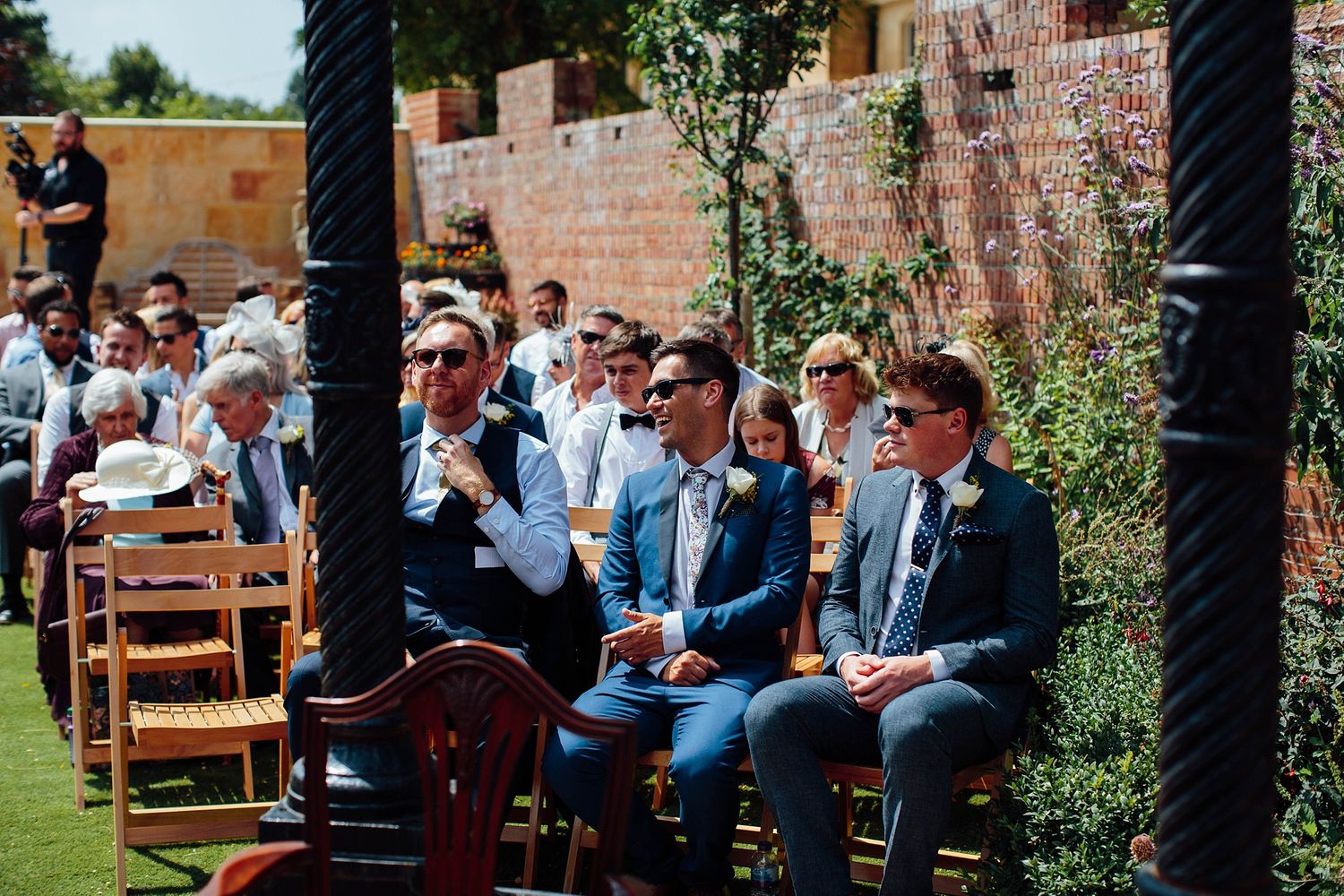 Wedding ceremony guests seated in wooden chairs among garden greenery and brick walls.