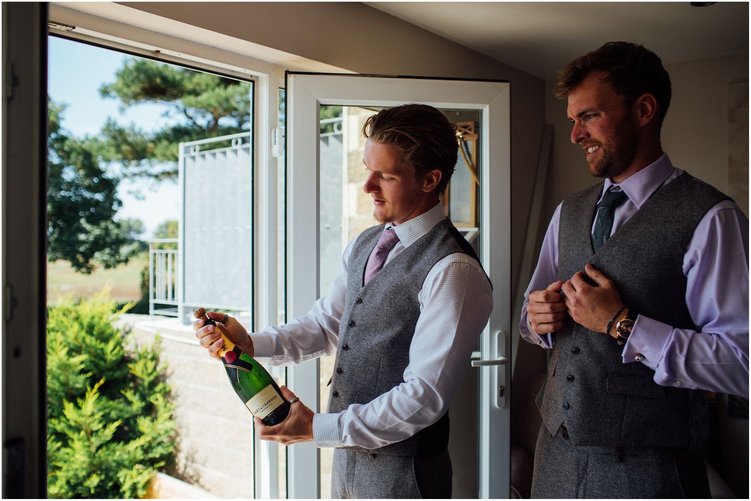 Two people in formal attire examine a bottle by large windows with natural light.