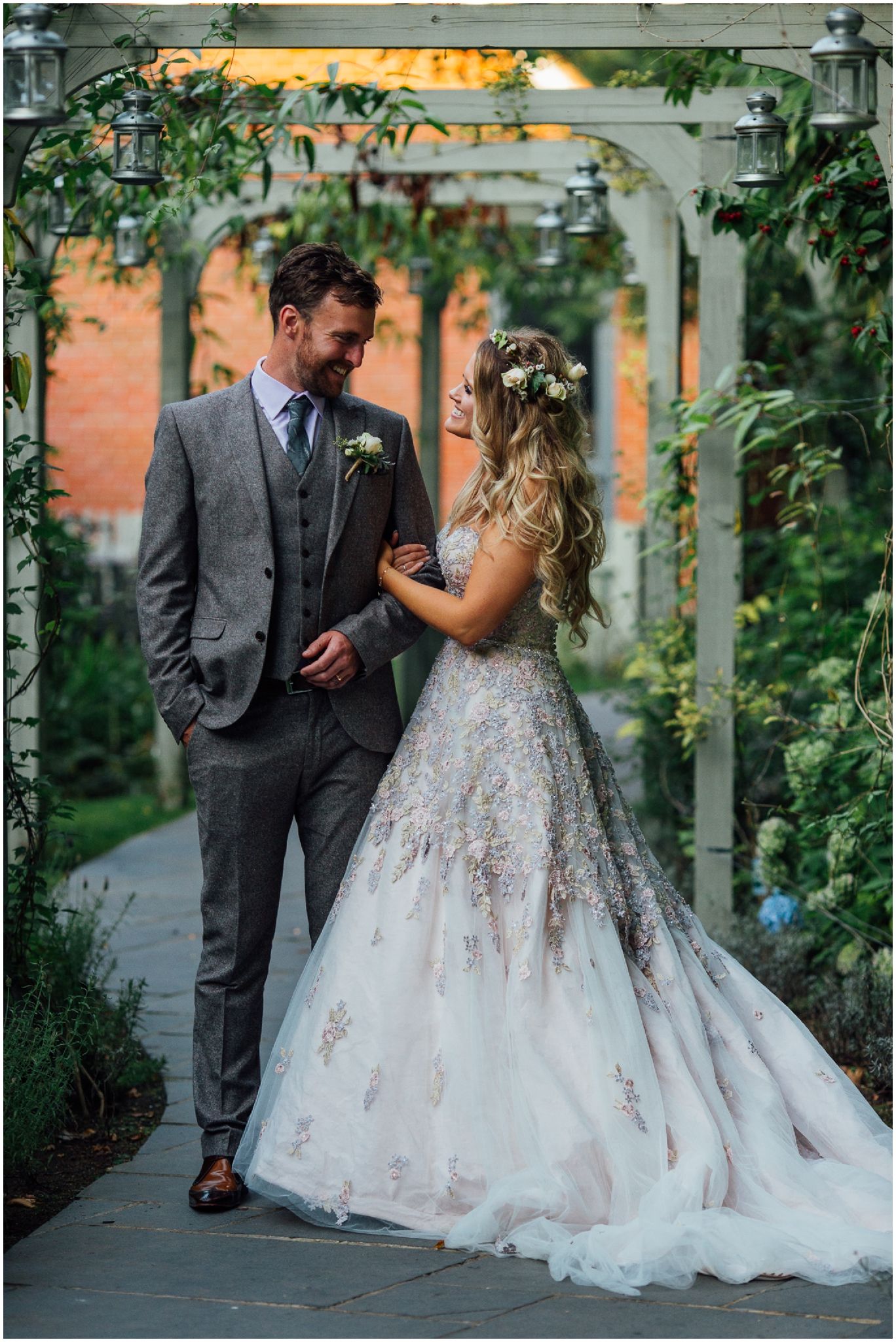 A romantic wedding couple poses on a garden walkway surrounded by greenery and flowers.