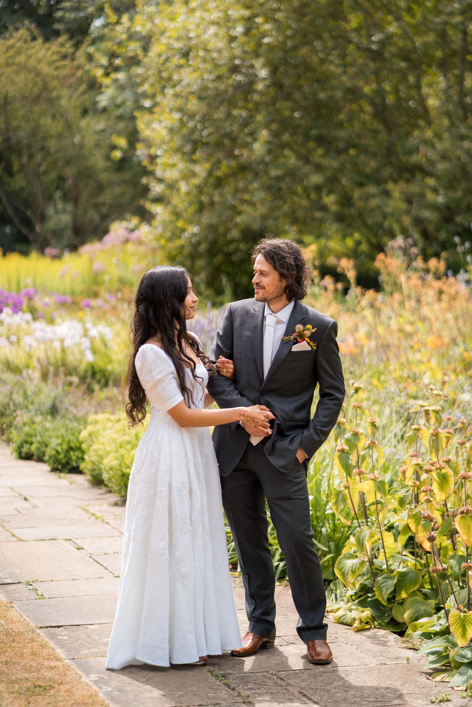 A bride and groom hold hands smiling in a lush garden, she in white dress, he in dark suit with boutonniere.