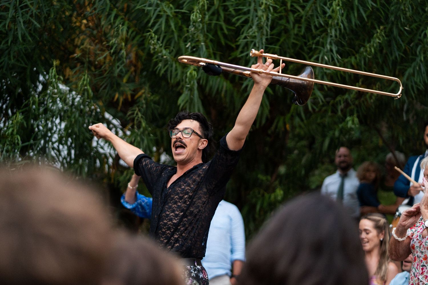 Energetic musician raises trombone overhead while entertaining a crowd at an outdoor wedding celebration.