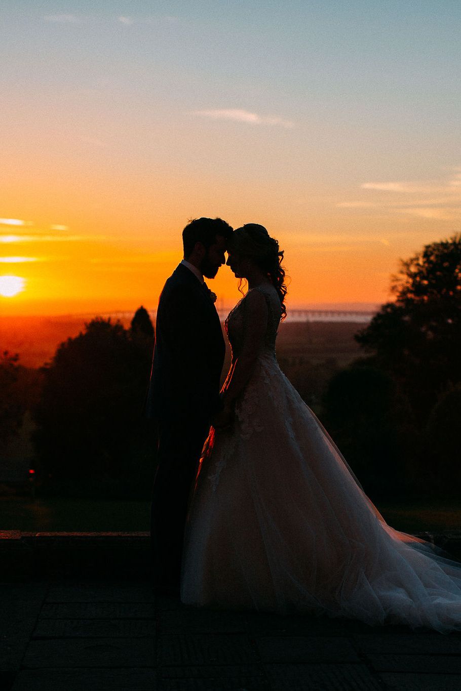 Silhouette of bride and groom facing each other at sunset with vibrant orange sky backdrop on wedding day.