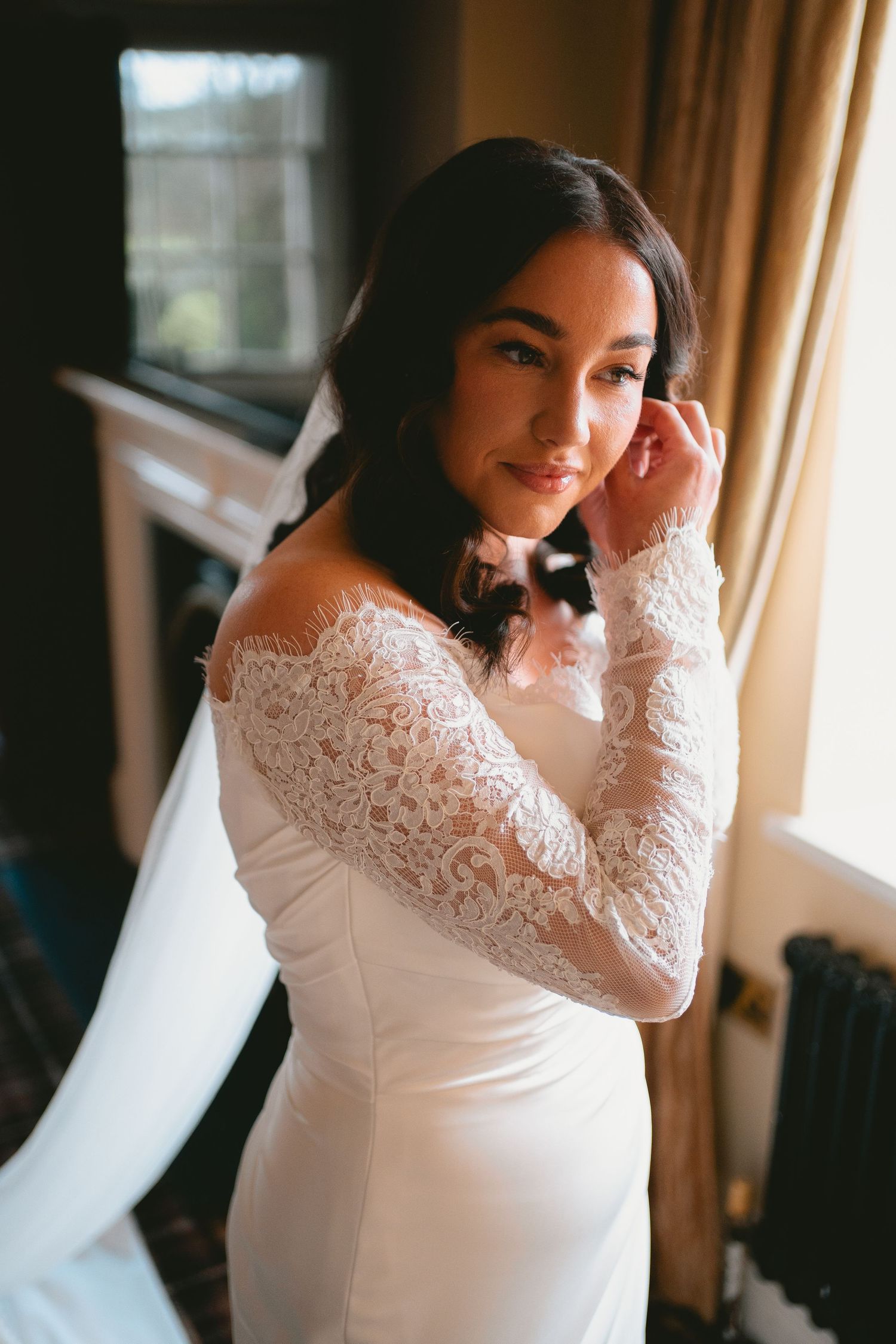 Bride in off-shoulder lace long-sleeve white wedding gown posing near a sunlit window, touching her ear.