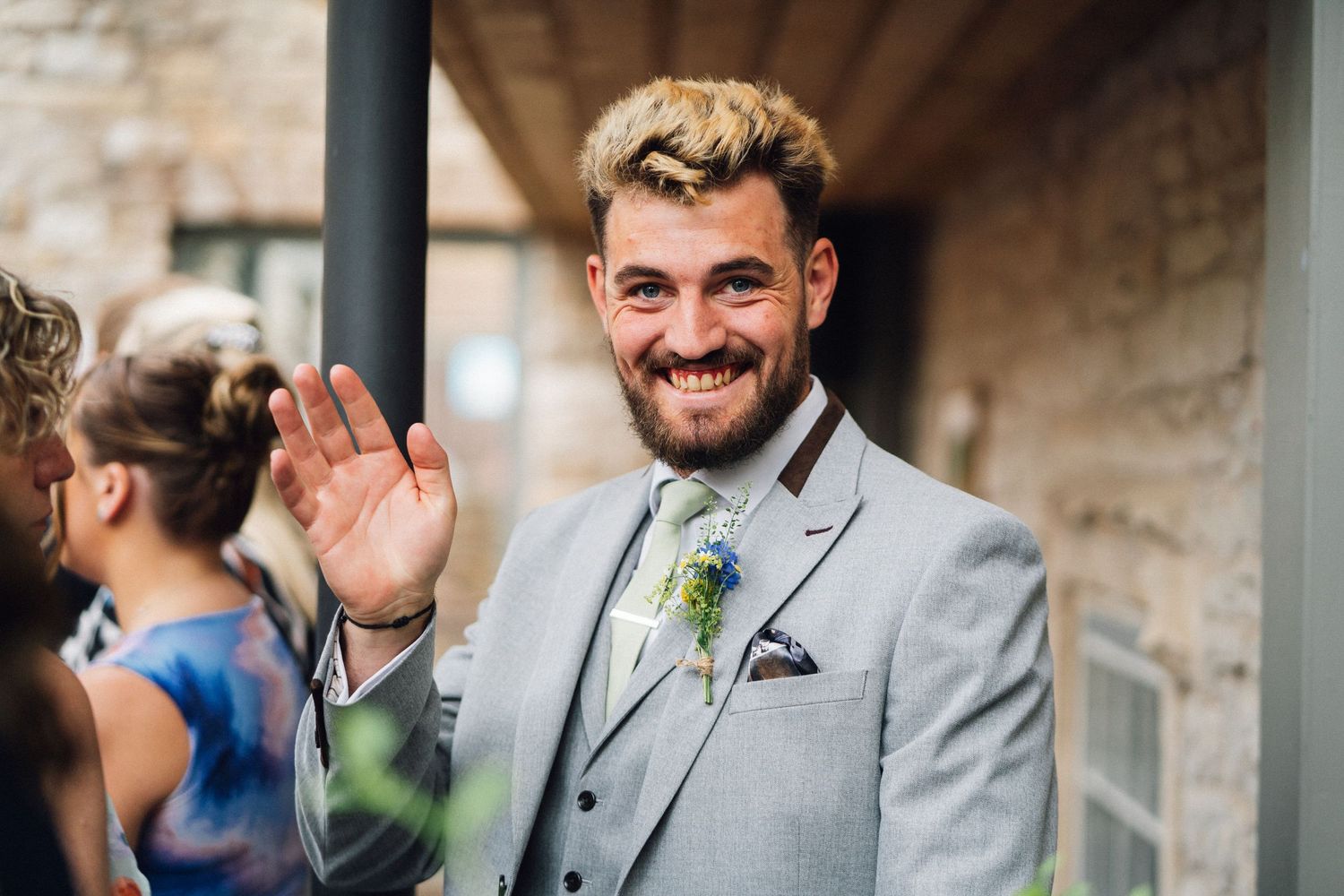 Smiling groom in grey suit with floral boutonniere waving at wedding celebration outdoors.