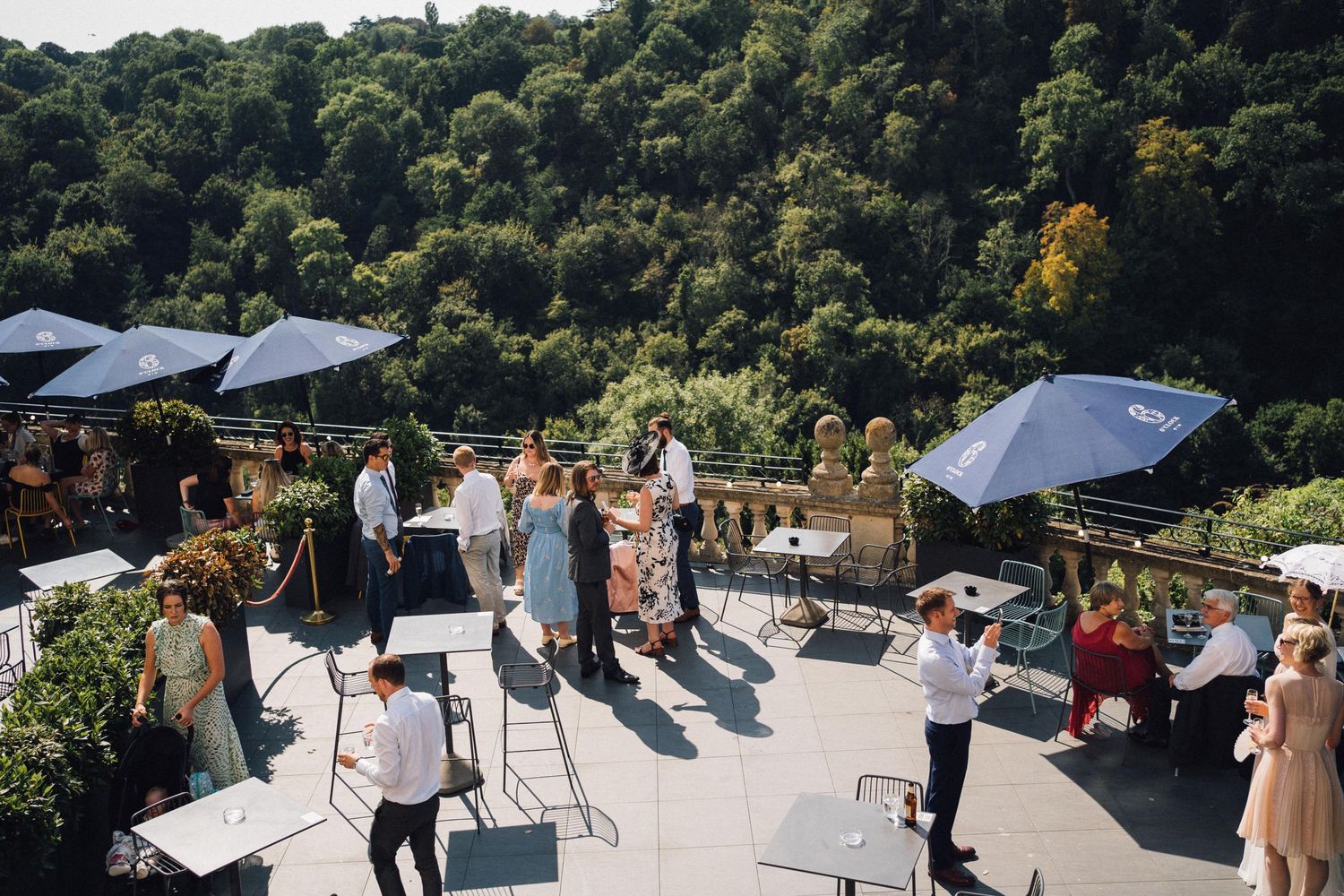 Elegant outdoor terrace wedding reception with guests mingling under blue umbrellas, lush green hillside backdrop.