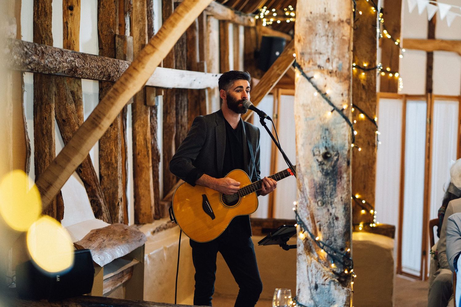 Musicians perform with acoustic guitars inside a rustic barn venue decorated with string lights and wooden beams.