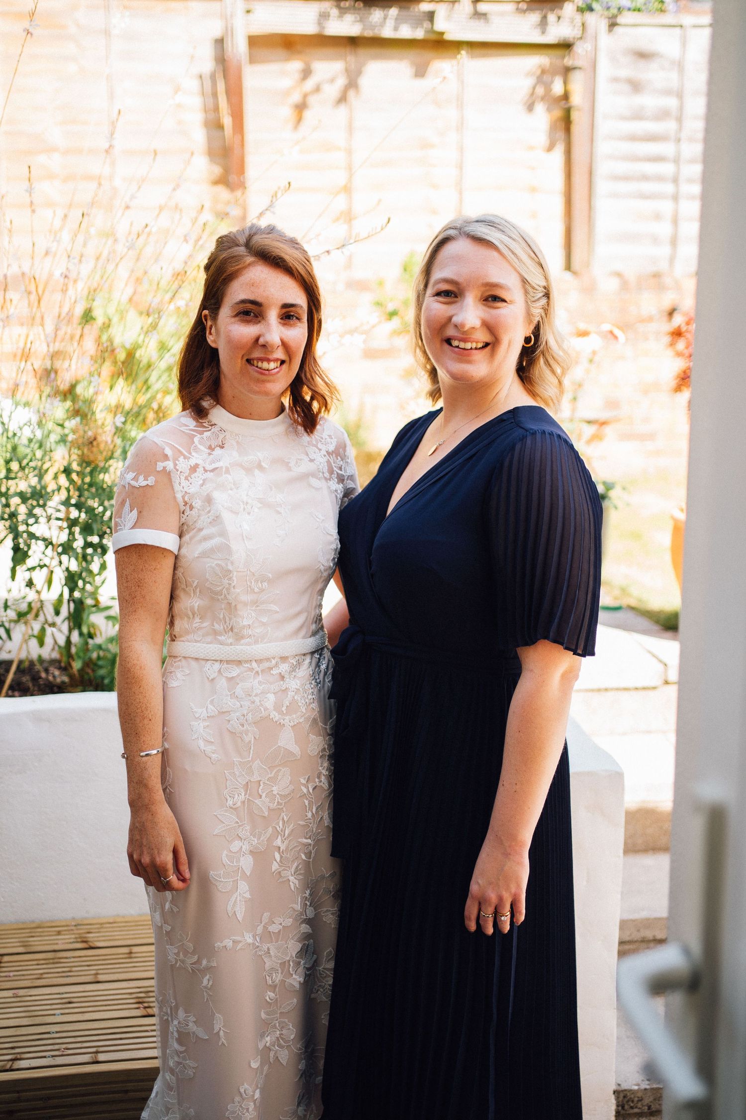 Two women pose together at an event, one wearing an elegant lace gown and the other in a navy blue dress.