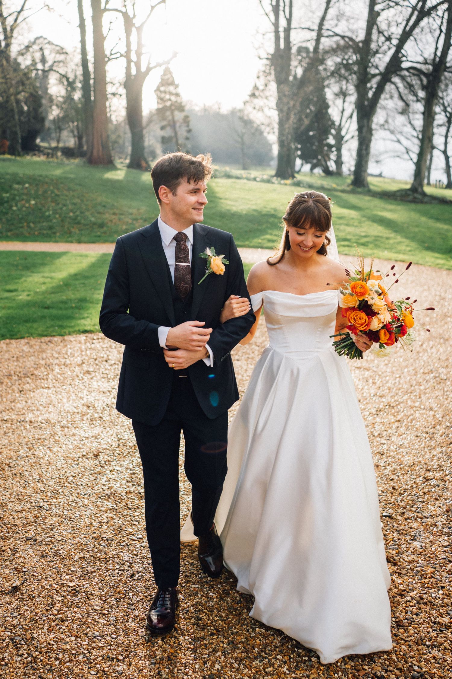 A newlywed couple walks along a tree-lined path with autumn-colored bouquet and elegant attire.
