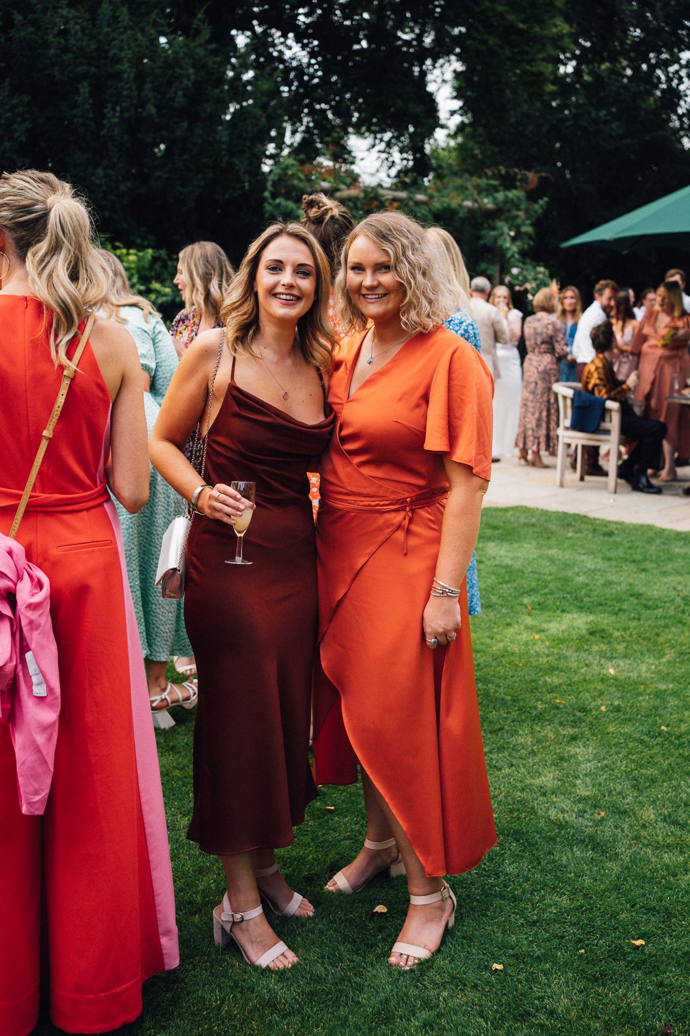 Bridesmaids in vibrant orange and burgundy dresses gather on green lawn during outdoor wedding reception.