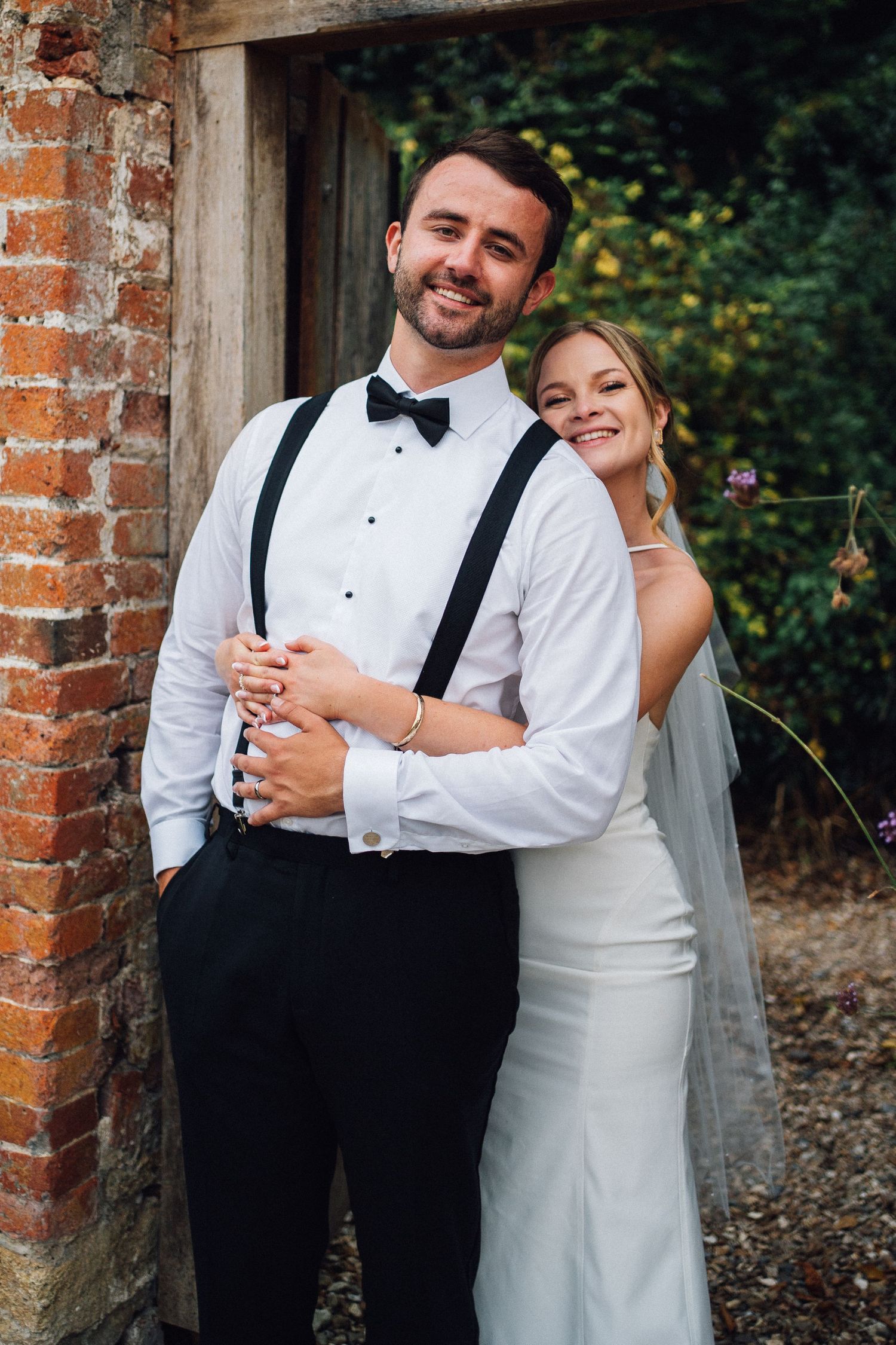 Couple in formal wedding attire pose playfully by brick archway.