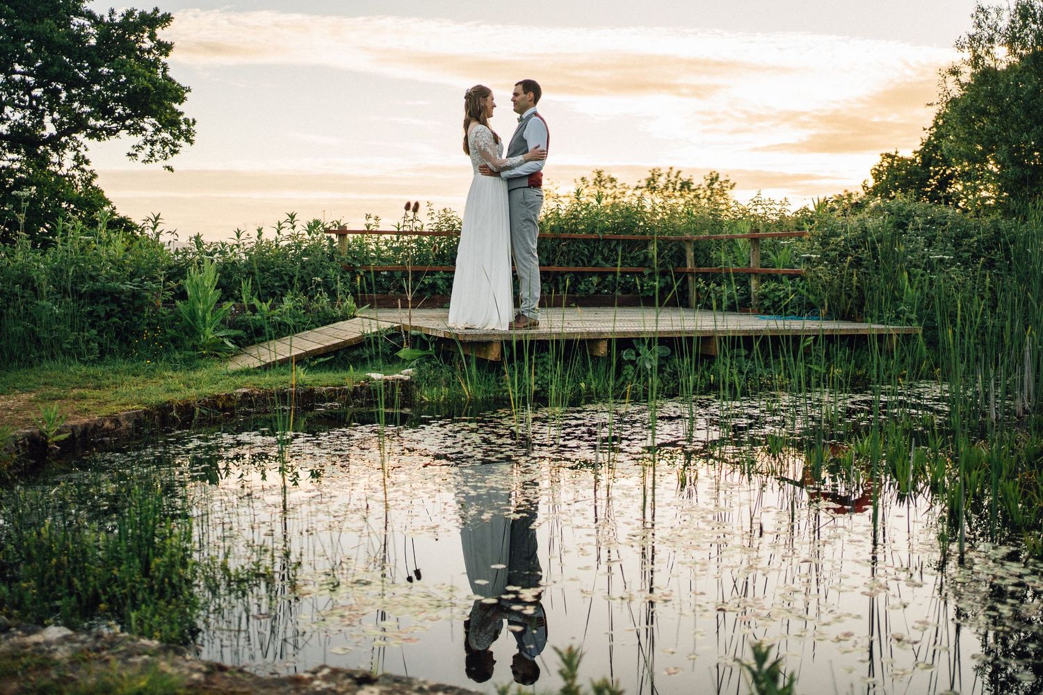 Wedding couple poses on wooden dock by tranquil pond during golden hour sunset.