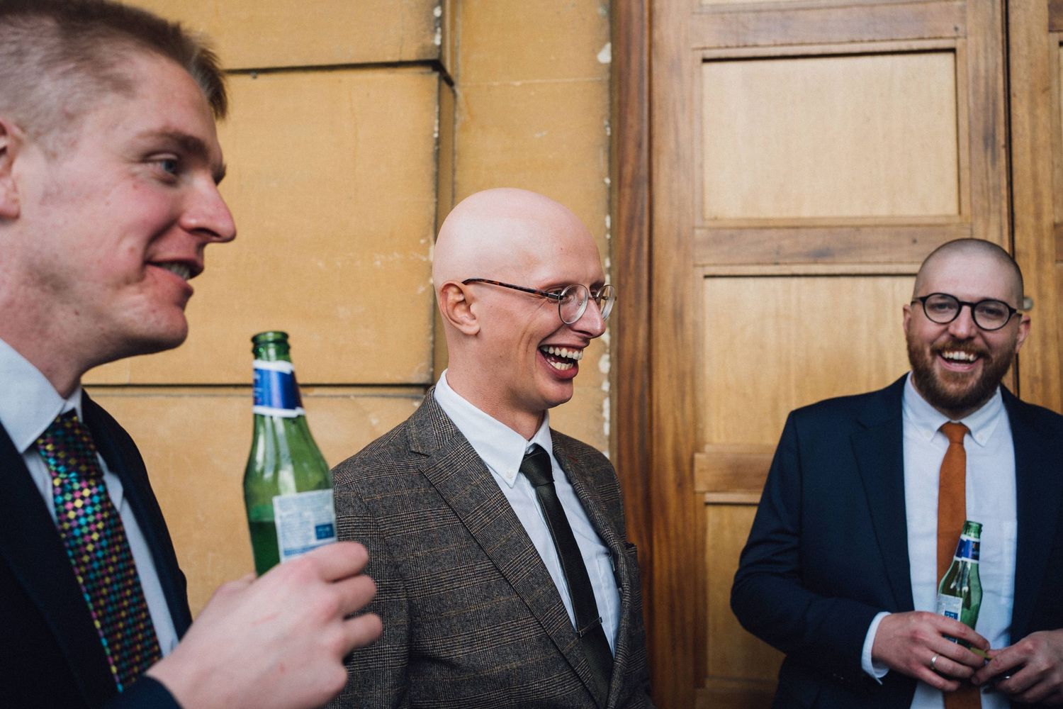 Wedding guests in formal attire share drinks and laughter during an indoor reception celebration.