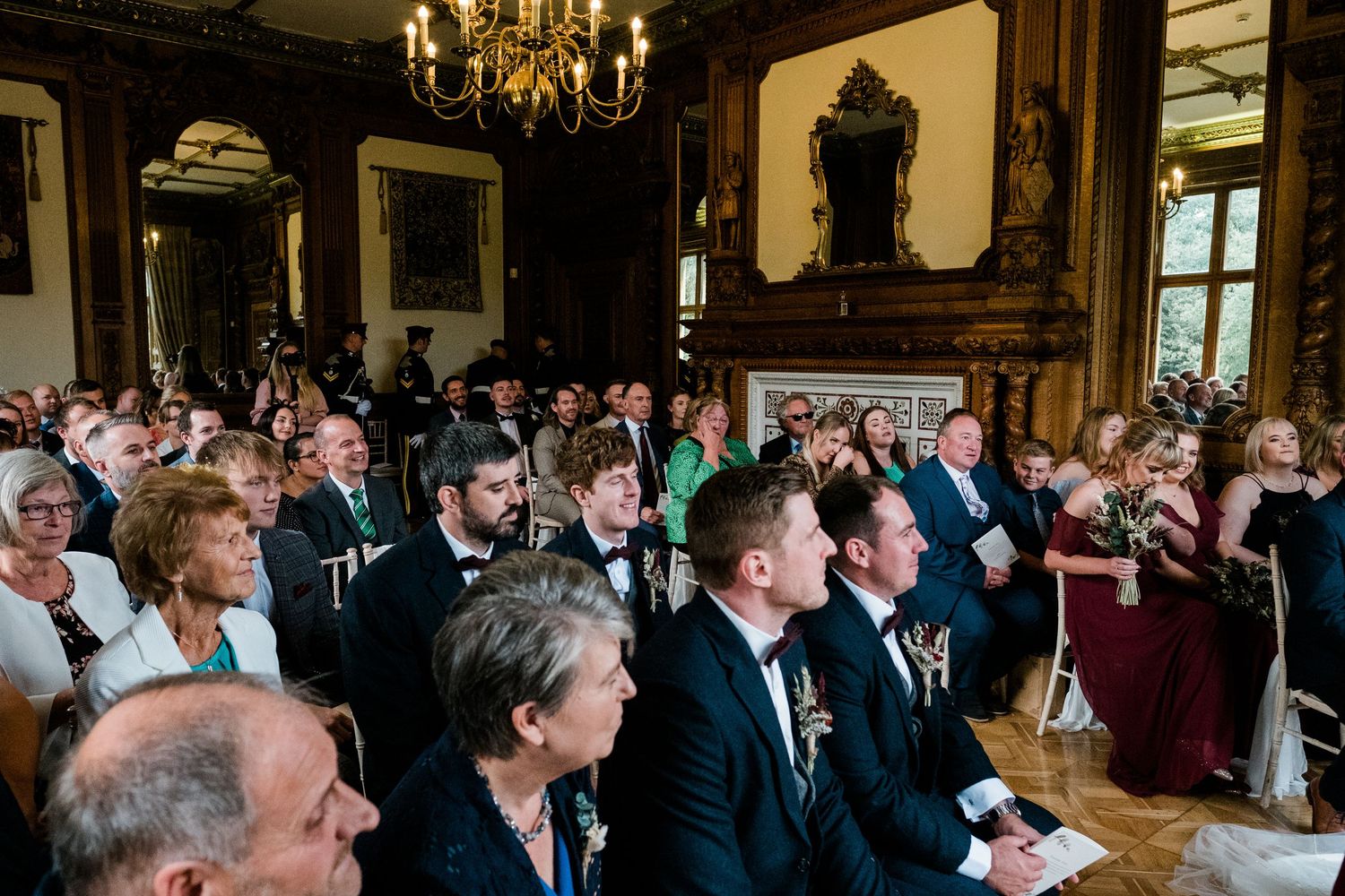 Wedding guests observe ceremony proceedings in an ornate room with chandelier and wood paneling.