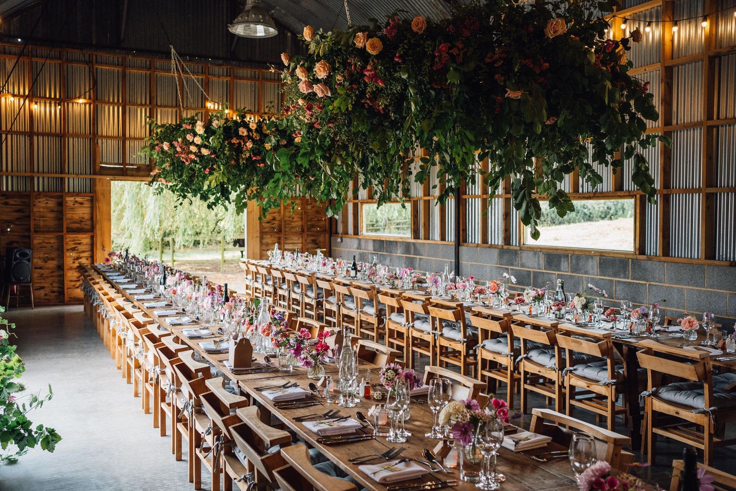 Elegant barn wedding reception with long wooden tables, floral centerpieces, and a lush hanging greenery installation overhead.