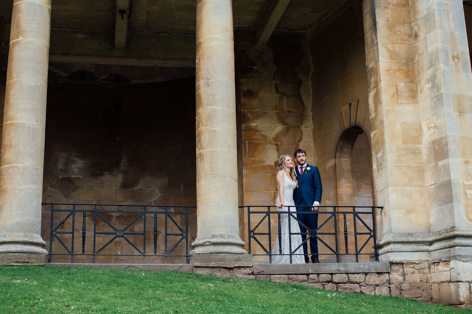 Bride and groom posing between stone columns on a balcony at a historic venue on their wedding day.